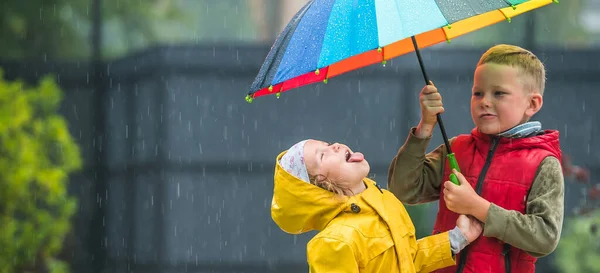 Маленькая девочка и мальчик с красочным зонтиком играют под дождем. Осенью дети играют на свежем воздухе в дождливую погоду. Осеннее веселье для детей. Малыш в плаще и сапогах ходит по саду — стоковое фото