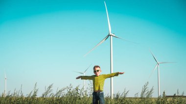 Çocuk rüzgar türbininin yanında rüzgarla oynuyor. Rüzgar değirmenleri. Elleri uçak gibi olan güneş gözlüklü çocuk. Yenilenebilir enerji ve sürdürülebilir kaynaklar - rüzgar değirmenleri