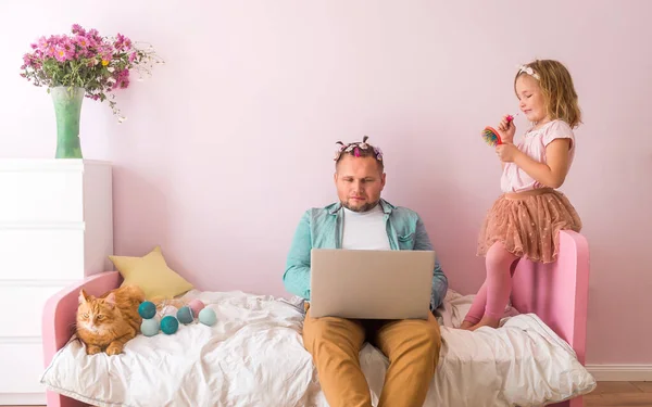 Work from home. Kid disturbing dad working remotely because of covid. Cute child girl putting on make up and make hair style. Man working on pink bed with laptop in girls room interior
