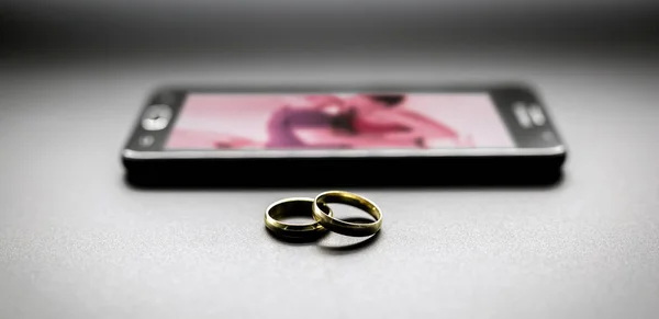 Concepto de amantes o traición conyugal, infidelidad. Pareja de anillos junto a un smartphone, con una imagen de una pareja borrosa besándose en la pantalla . — Foto de Stock