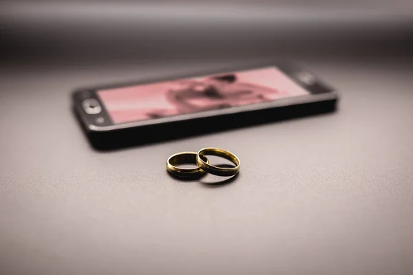 Concept d'amoureux ou de trahison conjugale, d'infidélité. Paire d'anneaux à côté d'un smartphone, avec une image d'un couple flou s'embrassant à l'écran . — Photo