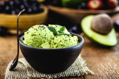 Arka planda tropikal meyveler bulunan avokadodan yapılmış vejetaryen dondurması el yapımı kil kasede servis edilir..