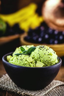 Arka planda tropikal meyveler bulunan avokadodan yapılmış vejetaryen dondurması el yapımı kil kasede servis edilir..