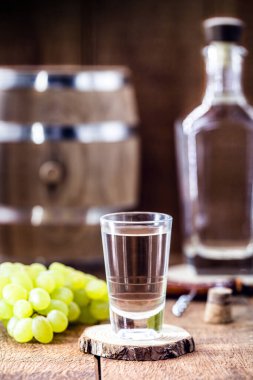Mayalanmış üzümlere dayanan damıtılmış içecek, shot bardağında, meşe fıçısıyla sade ahşap arka planda.