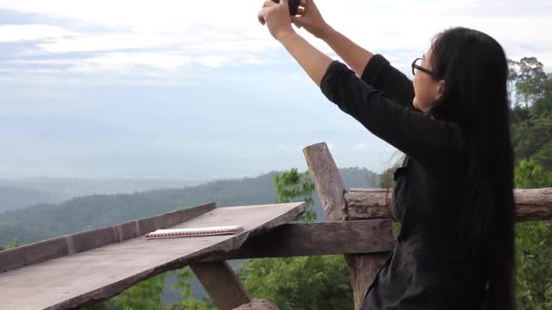 长黑头发亚洲妇女戴着眼镜与手机或智能手机在山林和天空视图背景的顶部 拍摄照片 — 图库视频影像