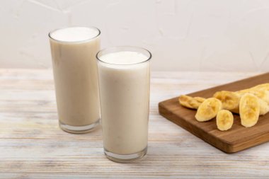 Muzlu milkshake. Serinletici muzlu smoothie, uzun bardaklarda sunuluyor, dilimlenmiş muzların yanında, ahşap panoda sergileniyor, iştah açıcı bir sahne yaratıyor. Muzlu smoothie.