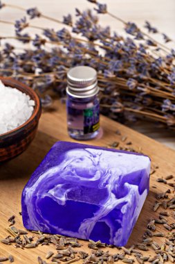 El yapımı sabun. Modelleme. Mor ve beyaz kıvrımlı lavanta sabun çubuğu ahşap yüzeyde duruyor. Yanında da esaslı yağ ve deniz tuzu var. Kurumuş lavanta çiçekleriyle çevrili. Sakinleştirici bir ortam oluşturuyor.