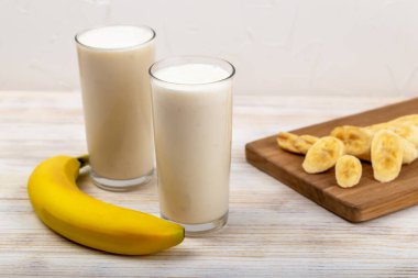 Muzlu milkshake. İki uzun bardakta sunulan taze muzlu smoothie yanında olgun muz ve tahta tahtada dilimler, sağlıklı ve lezzetli içecekler sergileniyor. Muzlu smoothie.