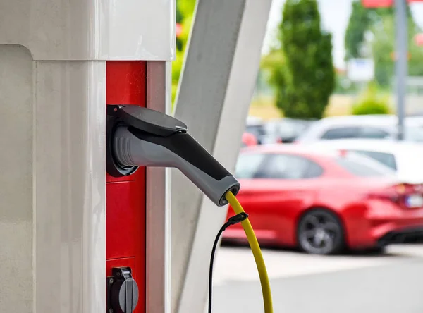 A charging cable for electric vehicles is plugged into a red-white charging station, cars are visible in the background