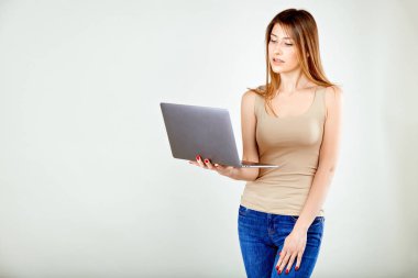 bej t-shirt ve mavi kot bir kız bir dizüstü bilgisayar tutan gri bir duvara karşı duruyor ve ilgi ile ekranına bakıyor