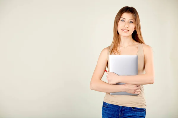 Ein Mädchen in beigem T-Shirt und blauer Jeans steht vor einer grauen Wand und hält einen geschlossenen Laptop in der Hand Stockfoto