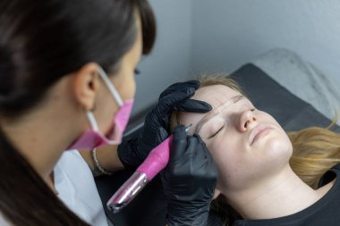 Yetenekli bir estetisyen, bir güzellik stüdyosunda rahat bir müşterinin kaşlarını titizlikle çizer. Kozmetik işlem sakin bir ortamda hassas araçlarla gerçekleştiriliyor.