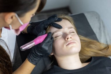 Bir güzellik uzmanı, tedavi yatağında yatan bir müşteriye kaş dövmesi yapar. Sanatçı modern bir salonda kaş bölgesini hassas aletlerle dikkatlice şekillendirir..