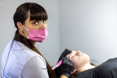 Maskeli bir güzellik teknisyeni, iyi aydınlatılmış bir stüdyodaki müşterilere mikropatinaj teknikleri uygular. Müşteri gözleri kapalı, mükemmel kaşlara odaklanır..