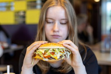 Parlak bir gündelik yemek alanında, genç bir kadın yoğun bir şekilde marul, domates ve peynir katmanlarıyla dolu büyük bir hamburgere odaklanır. Müşteriler yakınlarda konuştukça atmosfer canlanıyor..