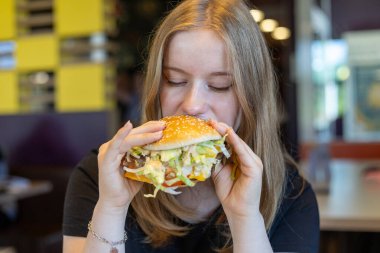 Genç bir kadın elinde marul, domates ve diğer malzemelerle dolu büyük bir burgerle masada oturuyor. Canlı bir restoran ortamında bir ısırık almaya hazır..