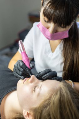 Yetenekli bir sanatçı, müşterisinin dudaklarına kalıcı makyaj uygulamaya odaklanır. Süreç, güzellik tedavi ortamında dikkatli bir teknik ve hassasiyet gösteriyor..