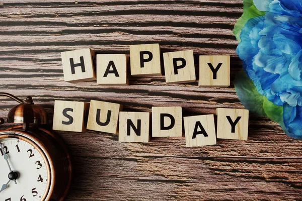 Alarm saati ve ahşap arka planda mavi çiçekli mutlu Pazar alfabesi mektupları.