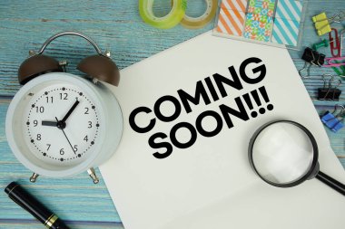 Coming Soon text on a paper surrounded by office supplies, a magnifying glass, and a clock on a blue wooden surface
