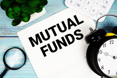 Mutual Funds written on paper placed on a blue wooden surface, alongside a magnifying glass, a clock, a calculator, and a small plant