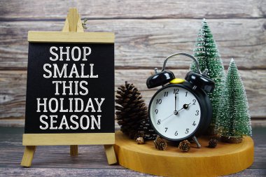 Festive holiday scene featuring a vintage alarm clock, pinecones, and miniature Christmas trees on a wooden base, with the text Shop Small This Holiday Season encouraging support for local businesses during the holiday season