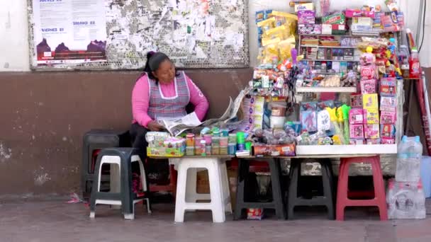 Cuenca, Εκουαδόρ - 20180920 - γυναίκα κάθεται από τον πίνακα των αγαθών προς πώληση, η ανάγνωση εφημερίδων ως πρόσωπο περπατά παρελθόν. — Αρχείο Βίντεο