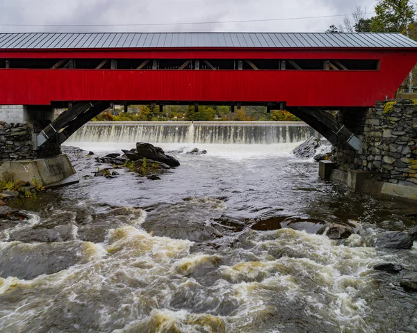 まず 1883 年に建てられた赤い屋根付きの橋にまたがる小さな水力発電所のダムで急速に流れる川 — ストック写真
