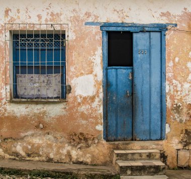 Yarı açık mavi kapı artı pencere. Kırık sarı duvarda taş basamaklar var.