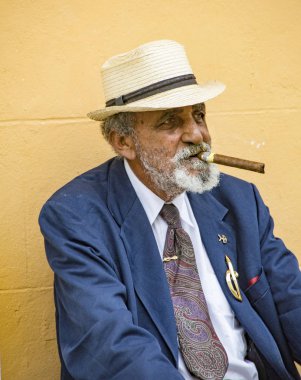 Adam bankta oturmuş, sarı bir duvara yaslanmış puro içiyor.