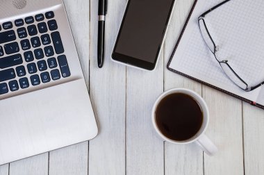 Bilgisayarlı, defterli, akıllı telefonlu beyaz masaüstünde bir fincan kahve.