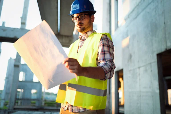 Portrait of male site contractor engineer with hard hat holding blue print paper at construction site,