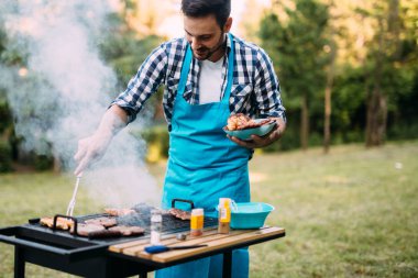 Handsome happy male preparing barbecue outdoors for friends clipart