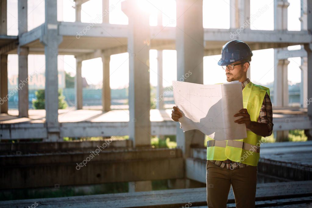 Portrait of male site contractor engineer with hard hat holding blue print paper at construction site,