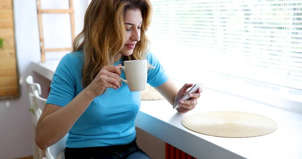 Beautiful woman using cell phone and drinking morning coffee at home