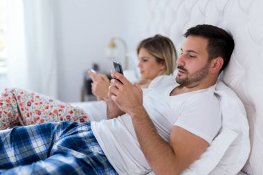 Telefonunu kullanarak yatak odasında sıkılmış genç çiftin resmi
