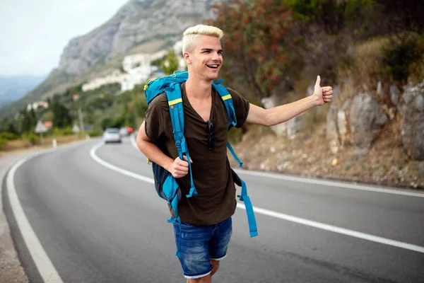 Hitchhiking young traveller man try to catch car on a forest road. World travel road trip, holiday hitch hike concept
