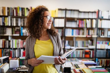 Eğitim, lise, üniversite, öğrenme ve insanların konsepti. Gülümseyen mutlu öğrenci kız kütüphanede kitap okuyor.