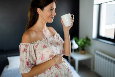 Genç, güzel, hamile bir kadın evinde çayını yudumluyor.