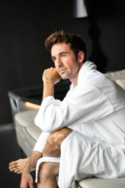 Young handsome man in the morning thinking while sitting in a hotel room in a robe clipart