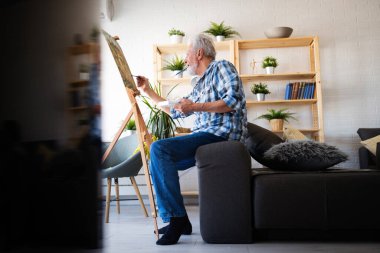 Evde eğlenmek için tuvale resim çizen mutlu emekli adam.