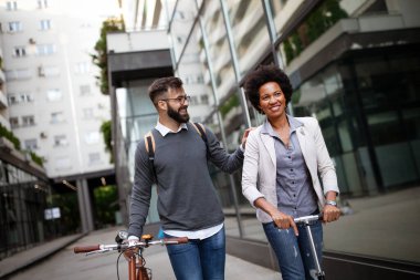 İki gülümseyen iş adamı elektrikli scooter kullanıyor, modern iş binasının önünde bisiklet sürüyor.