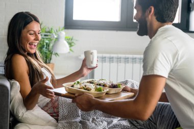 Güzel, genç, romantik bir erkek, bir kadına yatakta kahvaltı getirir ve uyandıktan sonra birlikte güler..