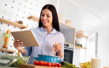 Mutfakta yemek hazırlarken bilgisayara bakan bir kadın. Güzel genç bir kadın yemek tarifi okuyor ya da internetten ders izliyor.