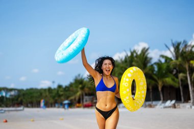 Cennette mutluluk. Gülümseyen genç bir kadın, tropikal kaçışının her anının tadını çıkarıyor. Plajda renkli şişirilebilir yüzüklerle oynuyor.
