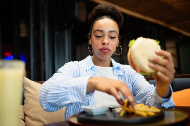A moment of pure happiness. Smiling young African American woman savoring her burger and fries at a cozy restaurant, embracing good food and positive vibes