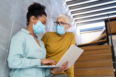 Teknoloji, yapay zeka, dijital yenilik ve salgın zamanlarında güvenli işyeri işbirliği için dizüstü bilgisayar kullanan maskeli kadın profesyoneller