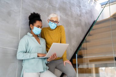 Teknoloji, yapay zeka, dijital yenilik ve salgın zamanlarında güvenli işyeri işbirliği için dizüstü bilgisayar kullanan maskeli kadın profesyoneller