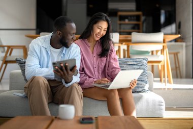 Genç ve çok ırklı çift evde bilgisayar ve dijital cihaz kullanırken eğleniyor. İnsanlar, teknoloji, online yaşam tarzı konsepti