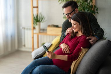 Hamilelik, teknoloji ve insan kavramı. Mutlu adam ve evde akıllı telefonları olan hamile karısı. Yeni mutlu aile.