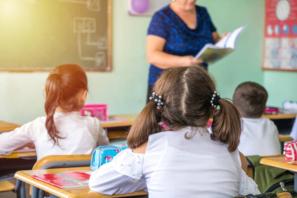 group of school kids sitting and listening to teacher in classroom from back.